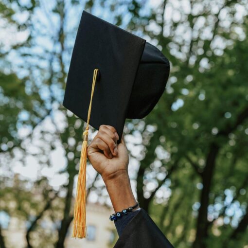 A person in a graduation gown holds up a graduation cap with a yellow tassel outdoors, with trees in the background.