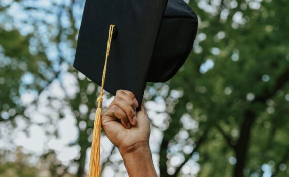 A person in a graduation gown holds up a graduation cap with a yellow tassel outdoors, with trees in the background.