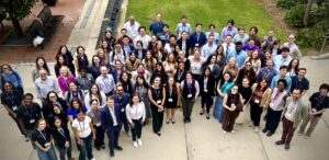 A large group of people stands outdoors on a paved walkway, looking up and smiling at the camera. They are wearing business casual attire and name badges.