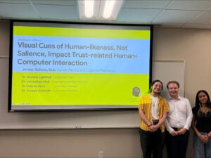Three people stand in front of a projected presentation slide titled "Visual Cues of Human-likeness, Not Salience, Impact Trust-related Human-Computer Interaction.