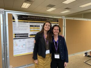 Two women stand and smile in front of a scientific research poster at a conference, wearing name badges and purple lanyards.