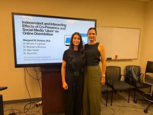 Two women stand side by side in a conference room. Behind them is a large screen displaying a research presentation on social media and online disinhibition.