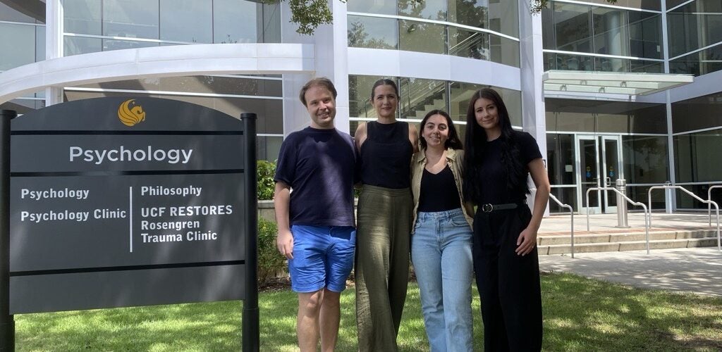 Four people stand in front of a building and sign for the UCF Psychology Clinic, Rosengren Trauma Clinic, and Philosophy department on a sunny day.