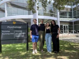 Four people stand in front of a building and sign for the UCF Psychology Clinic, Rosengren Trauma Clinic, and Philosophy department on a sunny day.