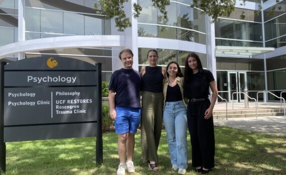 Four people stand in front of a building and sign for the UCF Psychology Clinic, Rosengren Trauma Clinic, and Philosophy department on a sunny day.