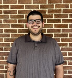 A man wearing glasses and a patterned polo shirt stands smiling in front of a brick wall, with a tattoo visible on his left forearm.