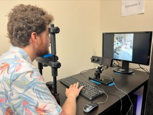 A man sits at a desk using an eye-tracking device while viewing a photo of a room and cat on a monitor. A keyboard, mouse, and labeled equipment are visible on the desk.