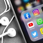 A smartphone screen displays icons for social media and messaging apps, including Facebook, Instagram, TikTok, YouTube, WhatsApp, Gmail, Twitter, Snapchat, and Telegram. Earphones are nearby.