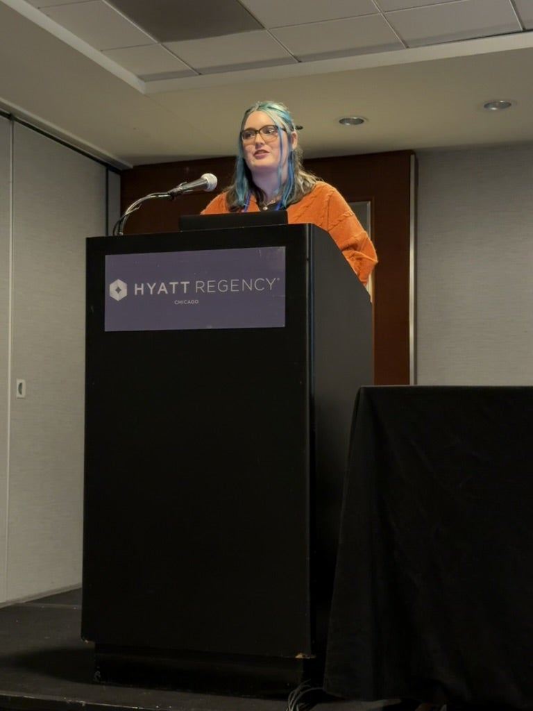 A person with blue hair and glasses speaks at a podium labeled "Hyatt Regency Chicago" in a conference room.