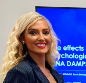 A woman with long blonde hair and a dark blazer smiles in front of a blue presentation screen with partial text visible.