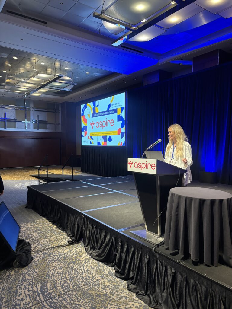 A person stands at a podium labeled "aspire," speaking on a stage in a conference room with a large screen displaying the Aspire logo and colorful graphics.