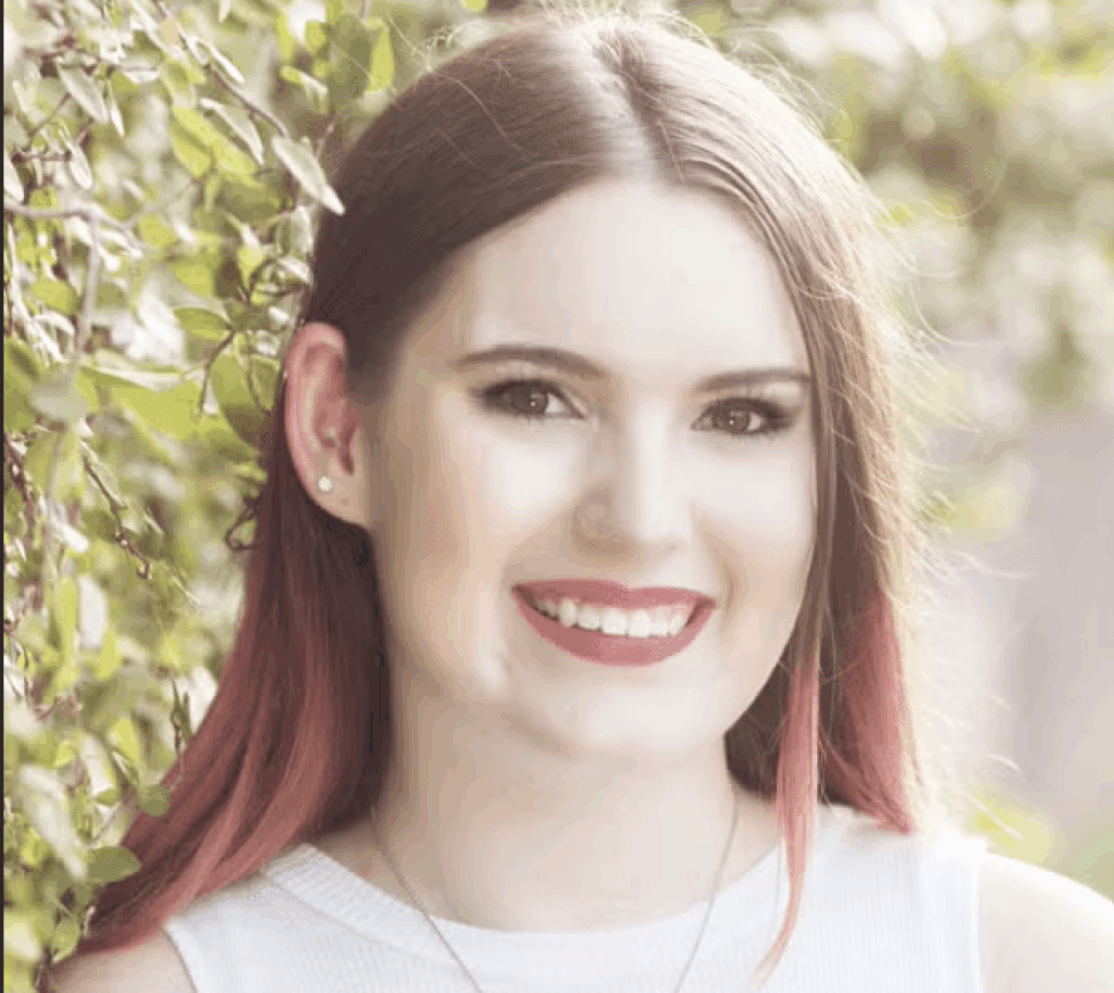 A young woman with long, straight brown hair and pink highlights smiles outdoors near leafy branches, wearing a sleeveless white top and stud earrings.