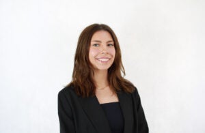 A woman with shoulder-length brown hair, wearing a black blazer and top, smiles at the camera against a plain white background.