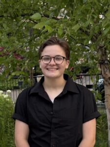 A person with short brown hair and glasses, wearing a black shirt, stands and smiles outdoors in front of green trees and plants.