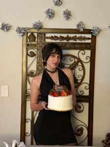 A person in a black dress holds a decorated cake with fruit and a "Black" sign, standing in front of an ornate metal gate and wall decorations.