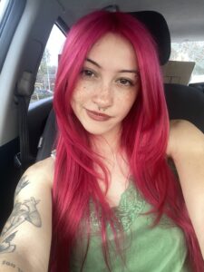 A person with long bright pink hair, a septum piercing, and visible tattoos sits in a car, wearing a green top and looking at the camera.