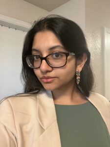 A woman with long dark hair and glasses wears a beige blazer, green top, and colorful dangling earring, standing indoors near a white door.