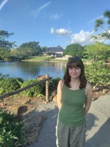 A woman in a green tank top stands on a gravel path in front of a pond, with trees, plants, and buildings visible in the background under a blue sky.