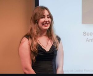 A woman with long brown hair wearing a black dress stands and smiles in front of a presentation screen.