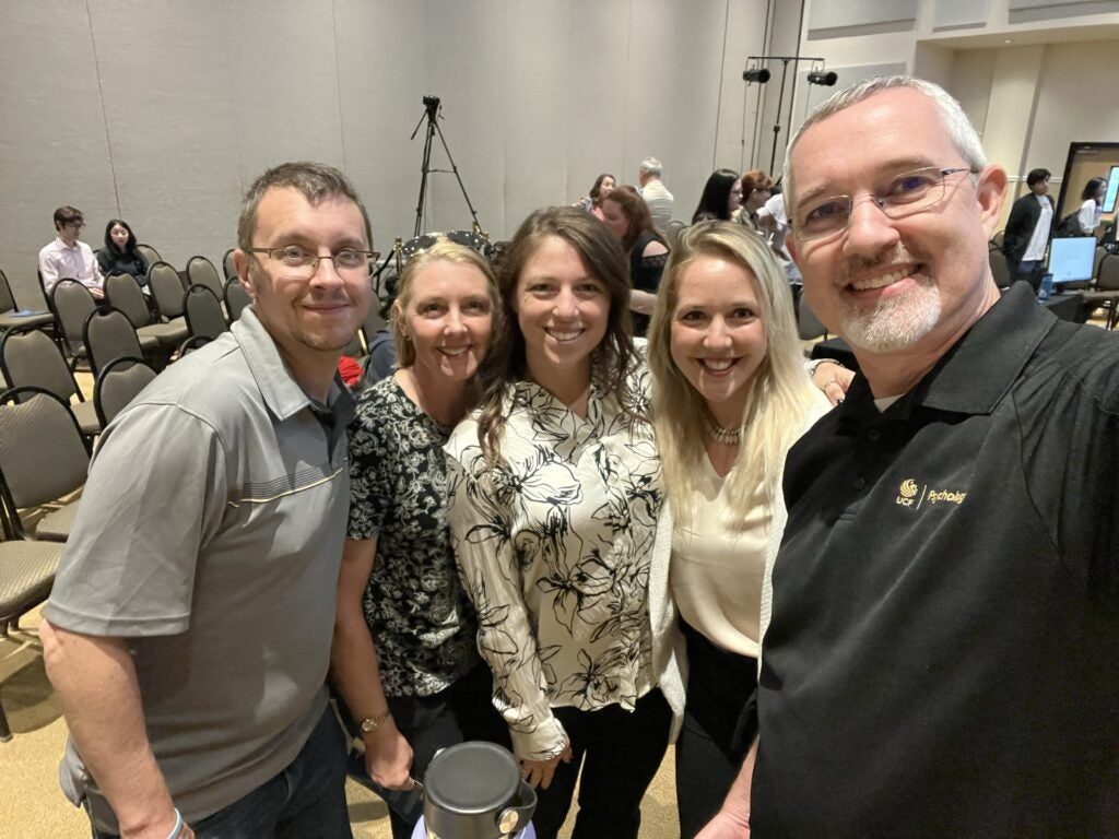 Five people smiling and posing for a group selfie in a conference room with rows of empty chairs in the background.