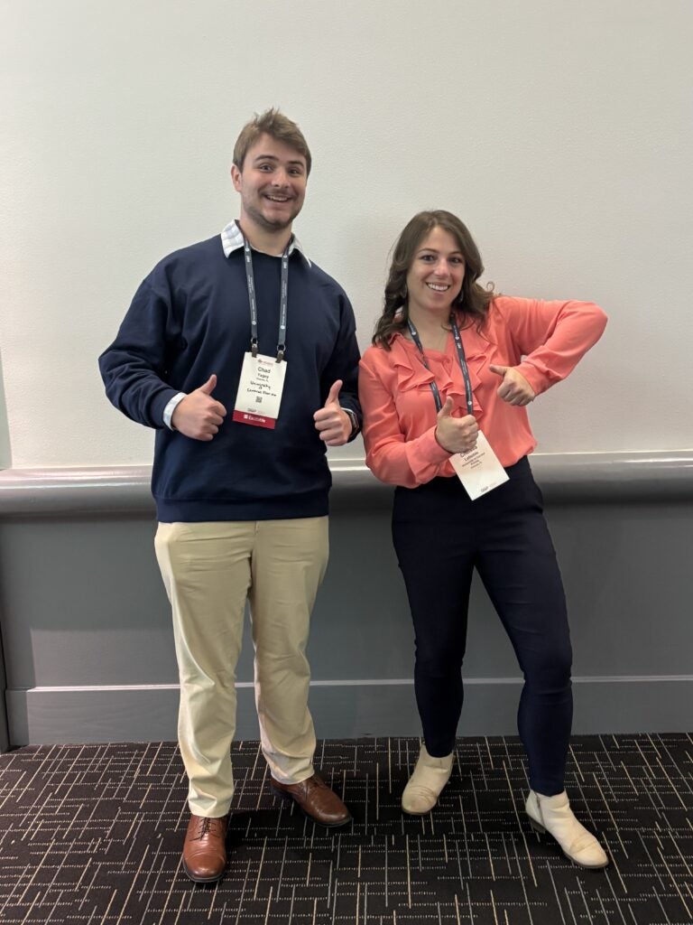 Two people stand side by side indoors, both smiling and giving thumbs up gestures, each wearing conference badges around their necks.