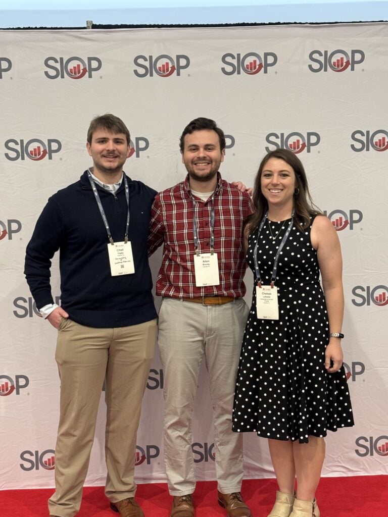 Three people stand side by side on a red carpet in front of a SIOP backdrop, wearing conference badges and business casual attire.