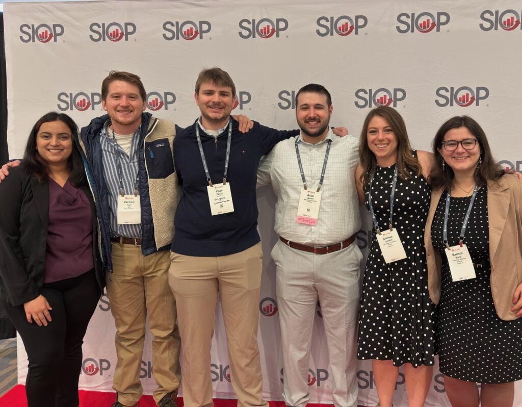 Six people standing in front of a SIOP-branded backdrop, smiling at the camera, wearing conference badges and business casual attire.