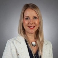 A woman with long blonde hair, wearing a white blazer and a dark top, poses in front of a plain grey background.