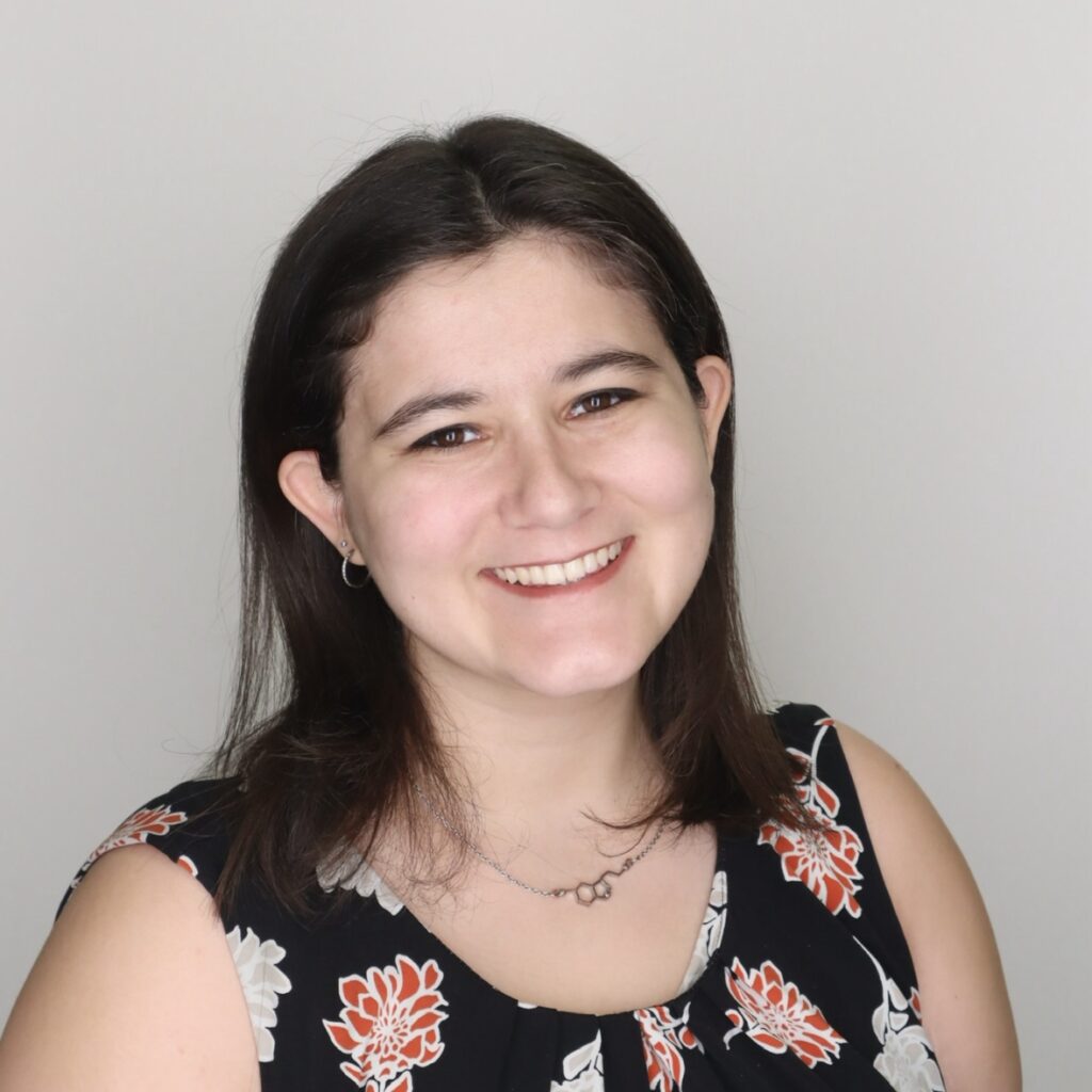 A woman with straight brown hair, wearing a black floral top and a necklace, smiles at the camera against a plain light background.