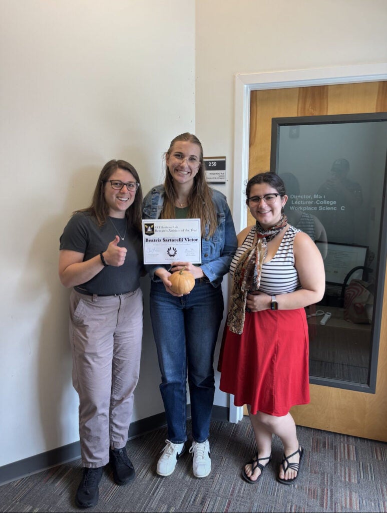 Three women stand indoors; one holds a certificate and a small pumpkin. Another gives a thumbs up. They are smiling, standing in front of an office door.