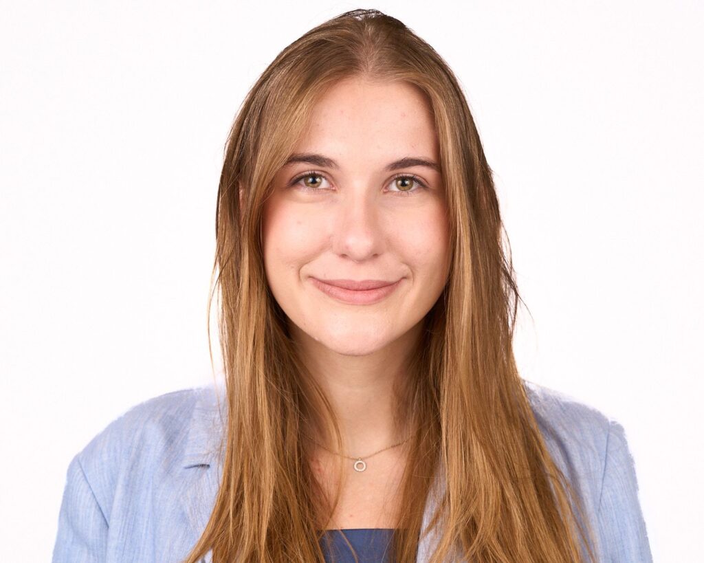 A young woman with long, straight light brown hair, wearing a light blue blazer and a necklace, smiles in front of a plain white background.