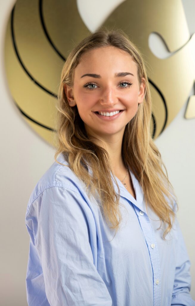 A young woman with long blonde hair, wearing a light blue shirt, smiles at the camera. A gold abstract logo is visible on the wall behind her.