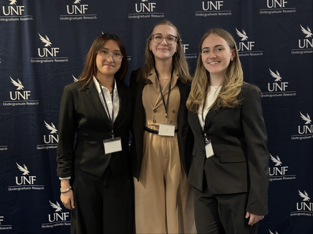 Three women wearing business attire stand and smile in front of a UNF Undergraduate Research backdrop, each wearing name badges on lanyards.