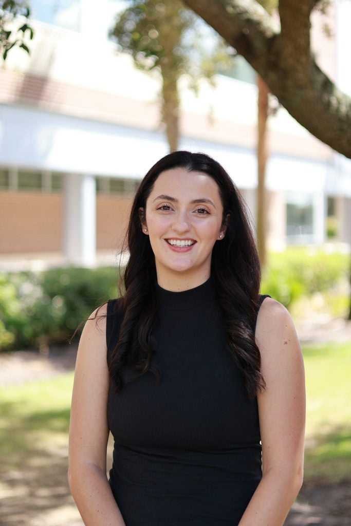 Woman with long dark hair in a sleeveless black top stands outdoors, smiling at the camera; trees and a building are in the background.