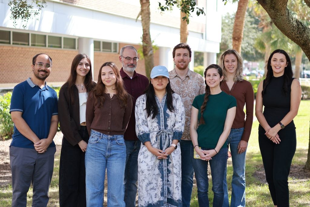 Nine people stand outdoors in front of a building, posing for a group photo on a sunny day with green grass and trees around them.