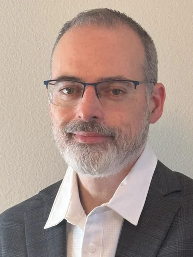 A man with short gray hair, a beard, and glasses wears a white collared shirt and a dark plaid suit jacket, standing in front of a plain light-colored wall.