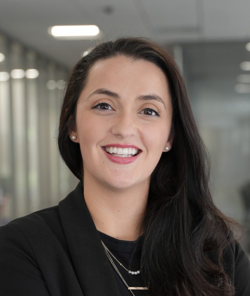 A woman with long dark hair wearing a black blazer and layered necklaces smiles in an office setting with glass walls and blurred background.