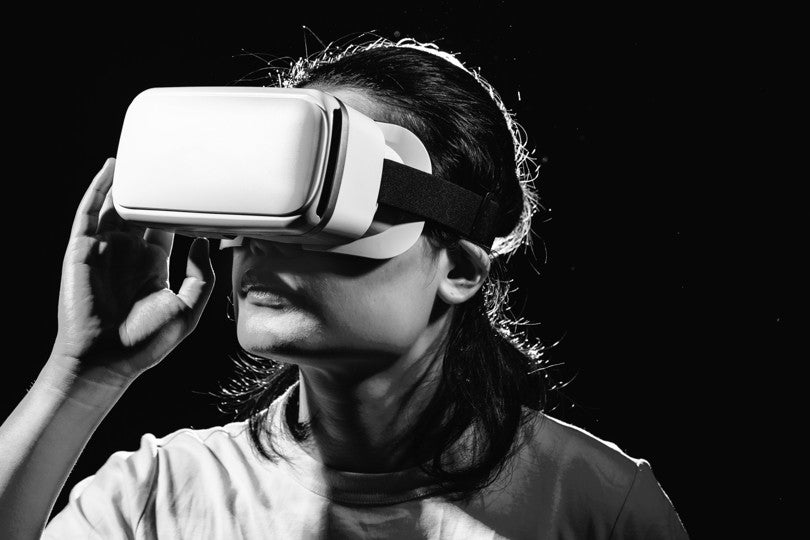 A person wearing a virtual reality headset looks upward, holding the device with one hand, against a dark background.