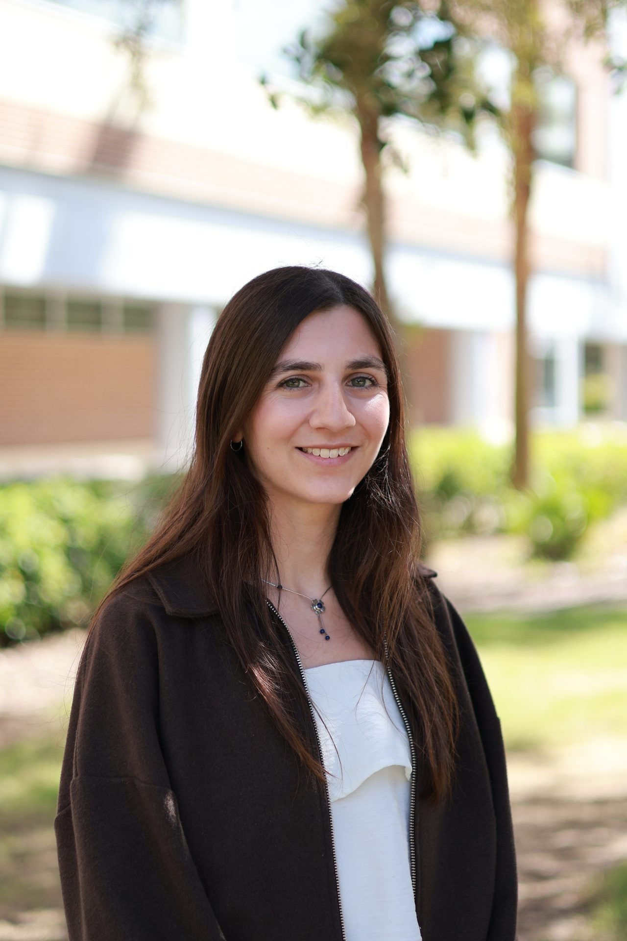 A young woman with long brown hair wearing a dark jacket and white top stands outside, smiling, with trees and a building in the blurred background.
