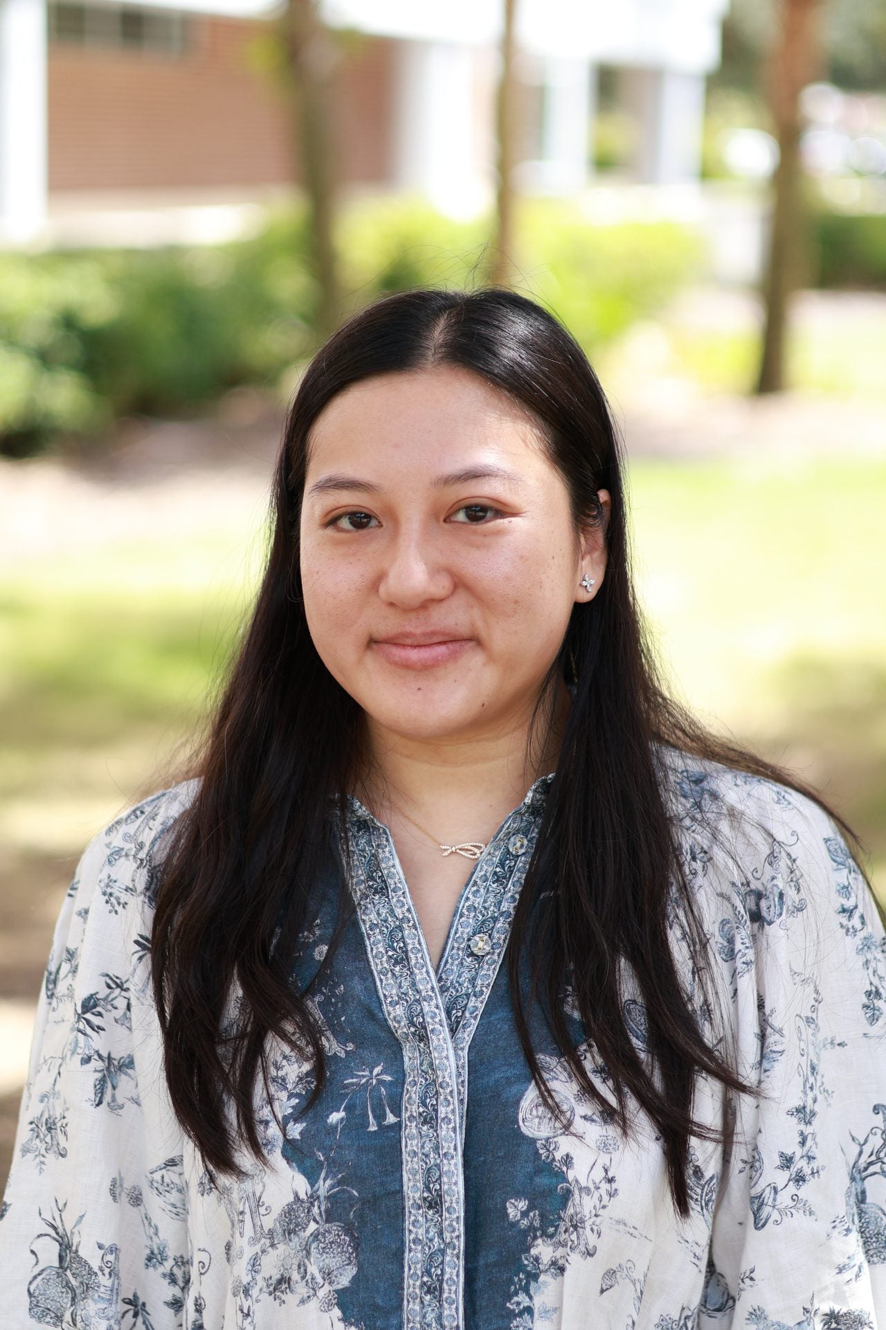 A woman with long dark hair stands outdoors, wearing a blue and white floral patterned top. The background is blurred with greenery and partial building visible.