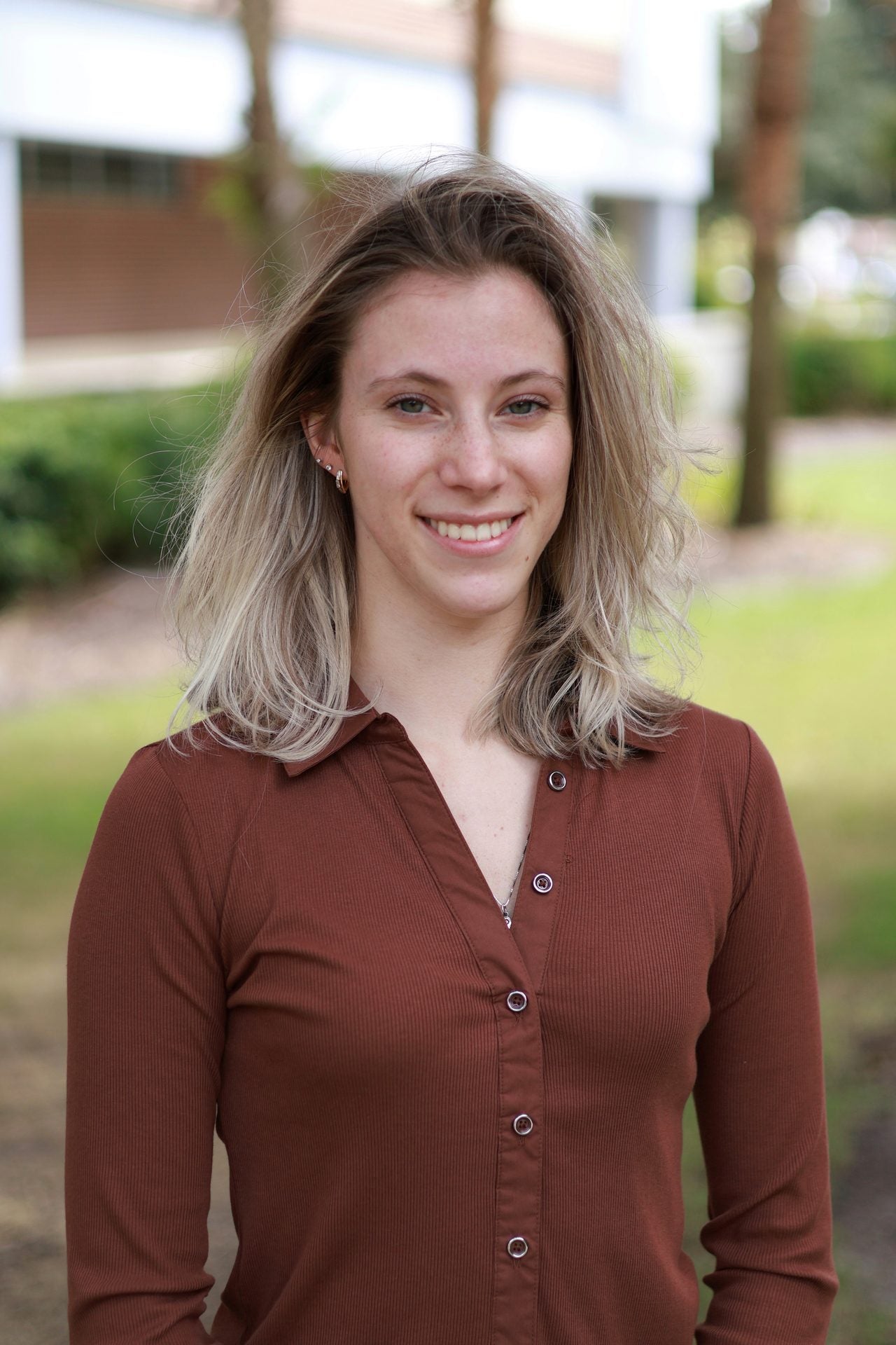 A woman with shoulder-length blonde hair wearing a brown button-up shirt stands outdoors, smiling at the camera with greenery and a building in the background.