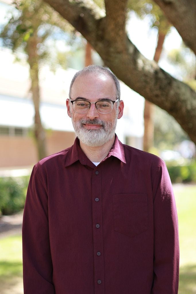 A man with glasses and a gray beard, wearing a maroon shirt, stands outdoors near trees on a sunny day.