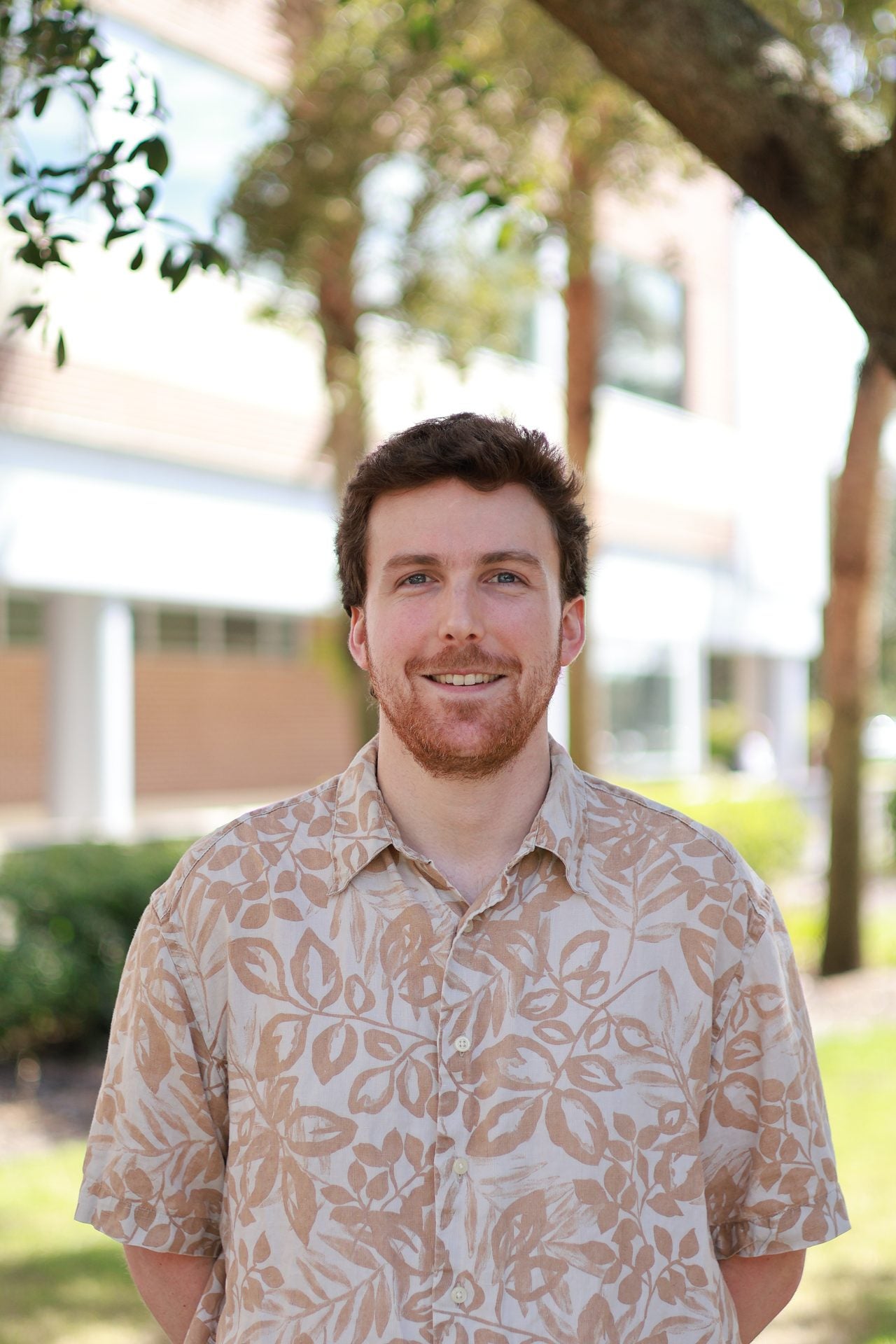 A man with brown hair and a beard wears a patterned short-sleeve shirt, standing outdoors with blurred trees and a building in the background.
