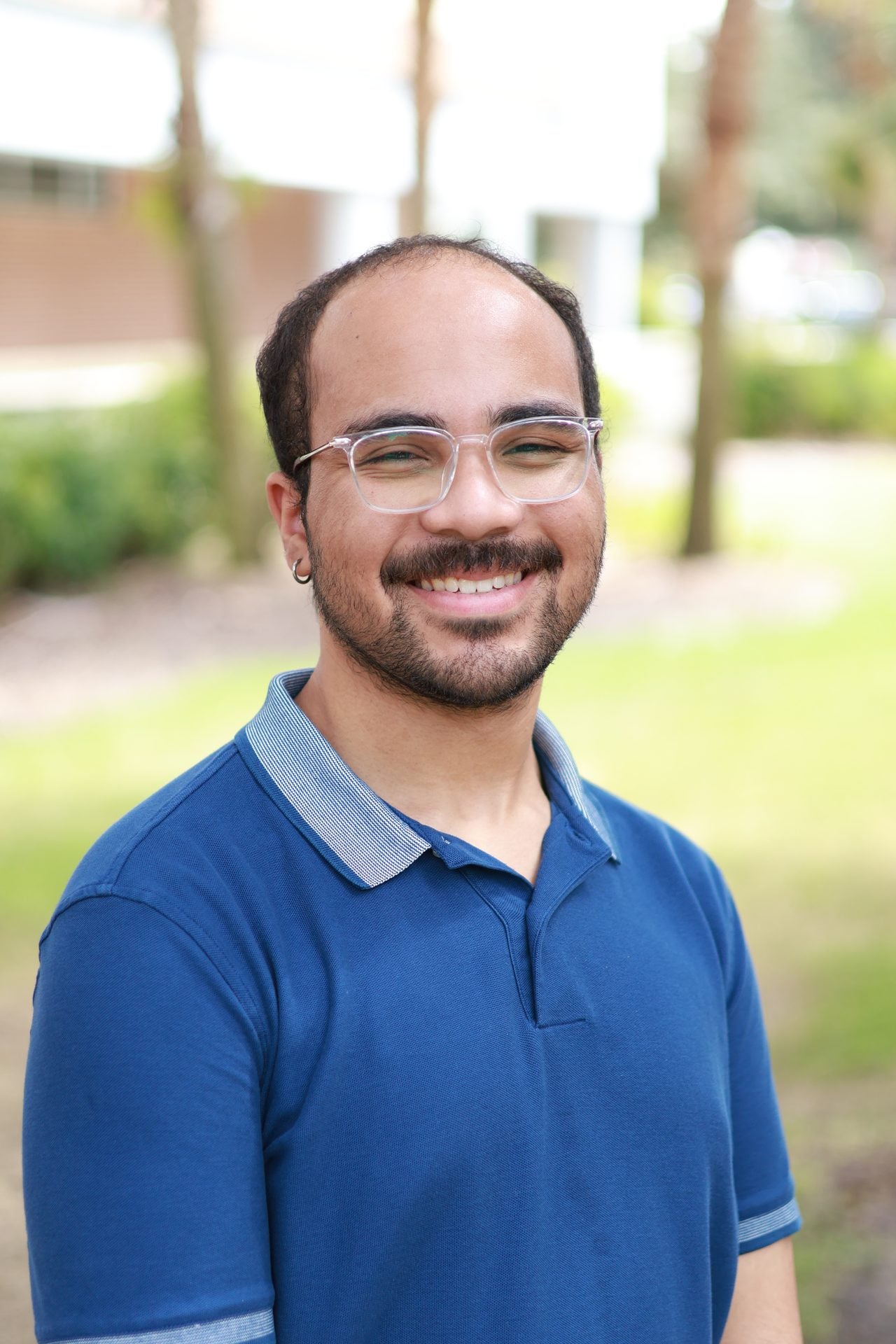 A person wearing glasses and a blue collared shirt stands outside, smiling at the camera with blurred greenery in the background.