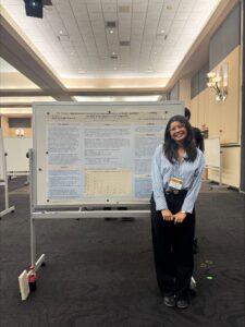 A woman stands and smiles in front of a large scientific research poster displayed on a whiteboard at a conference or academic event.