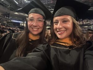 Two graduates wearing caps and gowns smile for a selfie at a crowded indoor graduation ceremony.