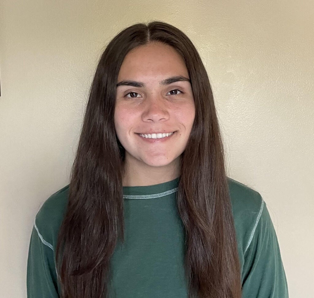 A smiling lab member with long brown hair and a green shirt stands in front of a plain beige wall, looking at the camera.