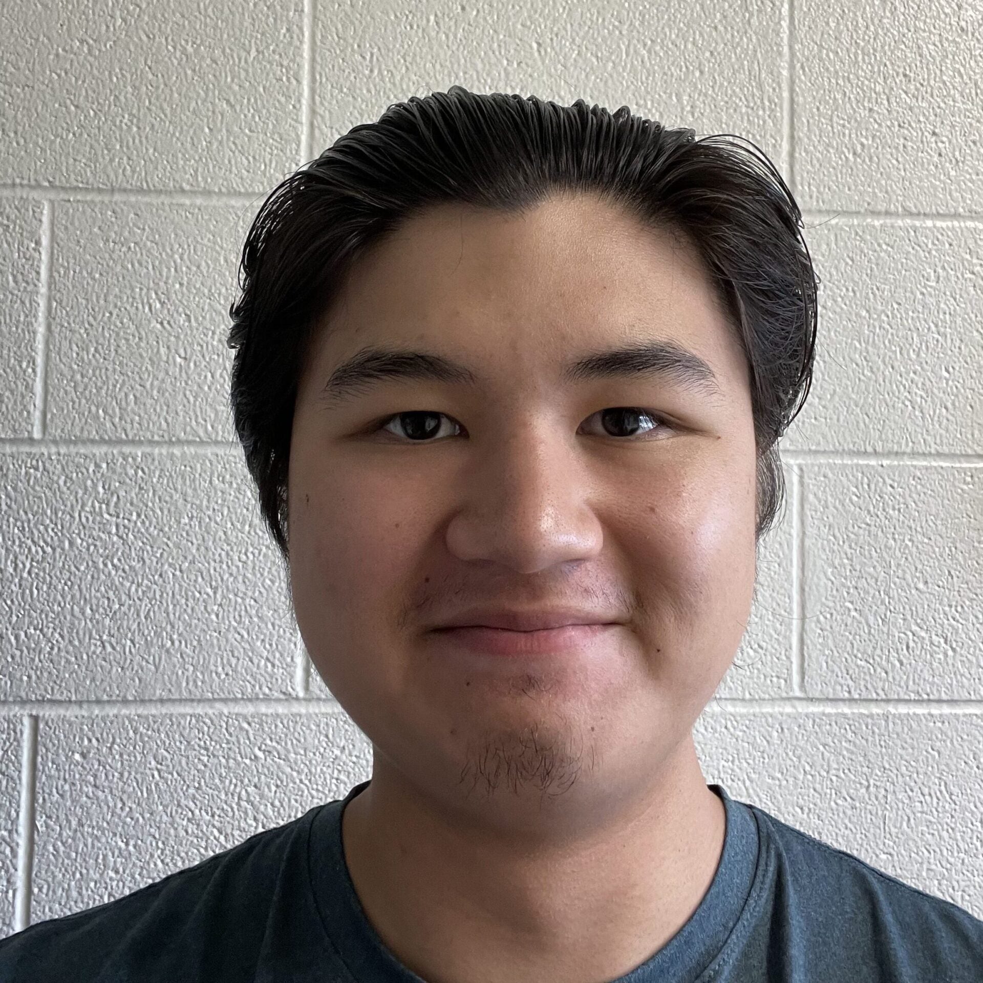 A lab member with short dark hair smiles slightly while standing in front of a white textured brick wall.