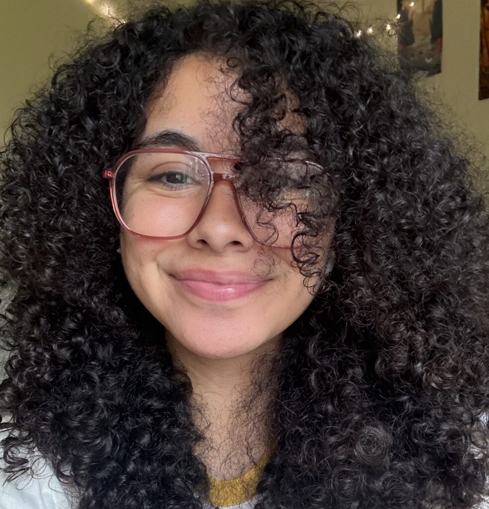 A lab member with curly hair and glasses smiles at the camera. String lights and posters are visible in the background.