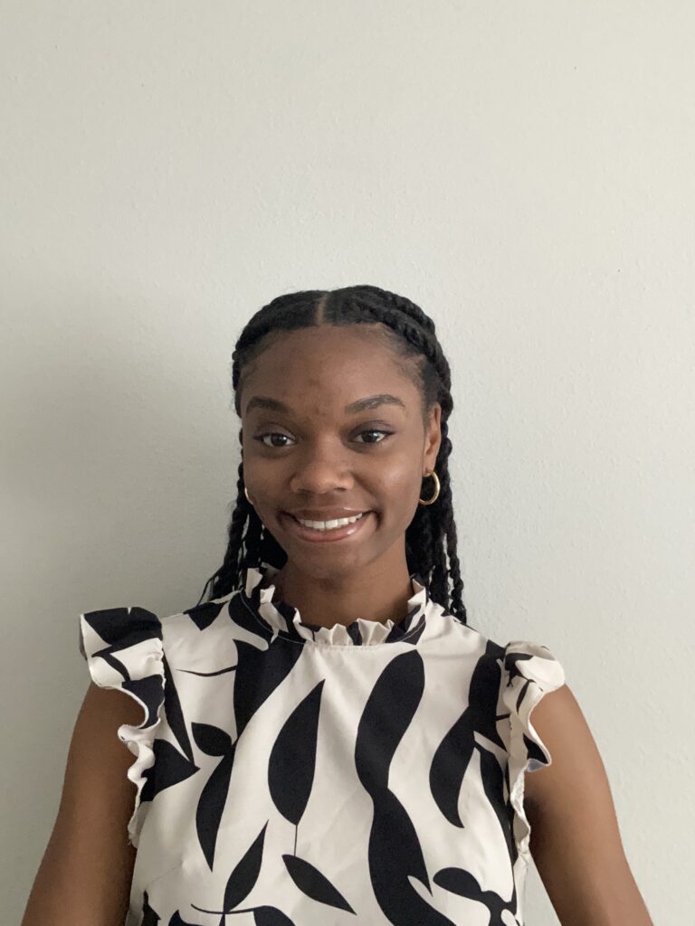 A woman with braided hair, one of the lab members, wears a sleeveless black-and-white patterned top and stands in front of a plain light-colored wall, smiling at the camera.